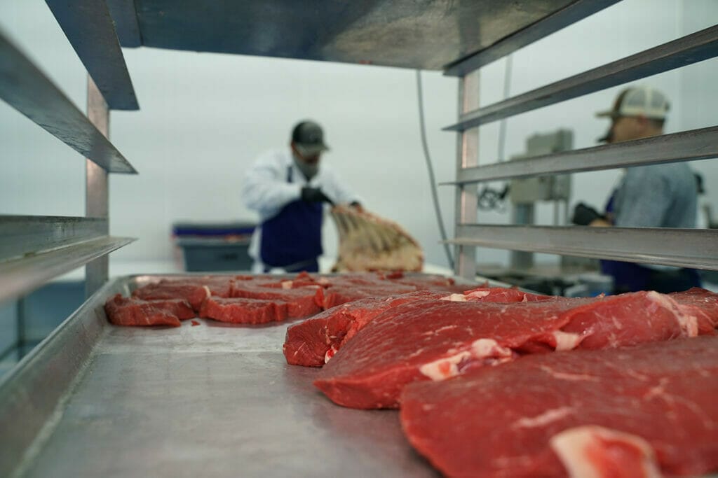 Picture of a butcher engaged in custom meat processing, viewed from the far side of a meat rack that is packed with beef steaks.